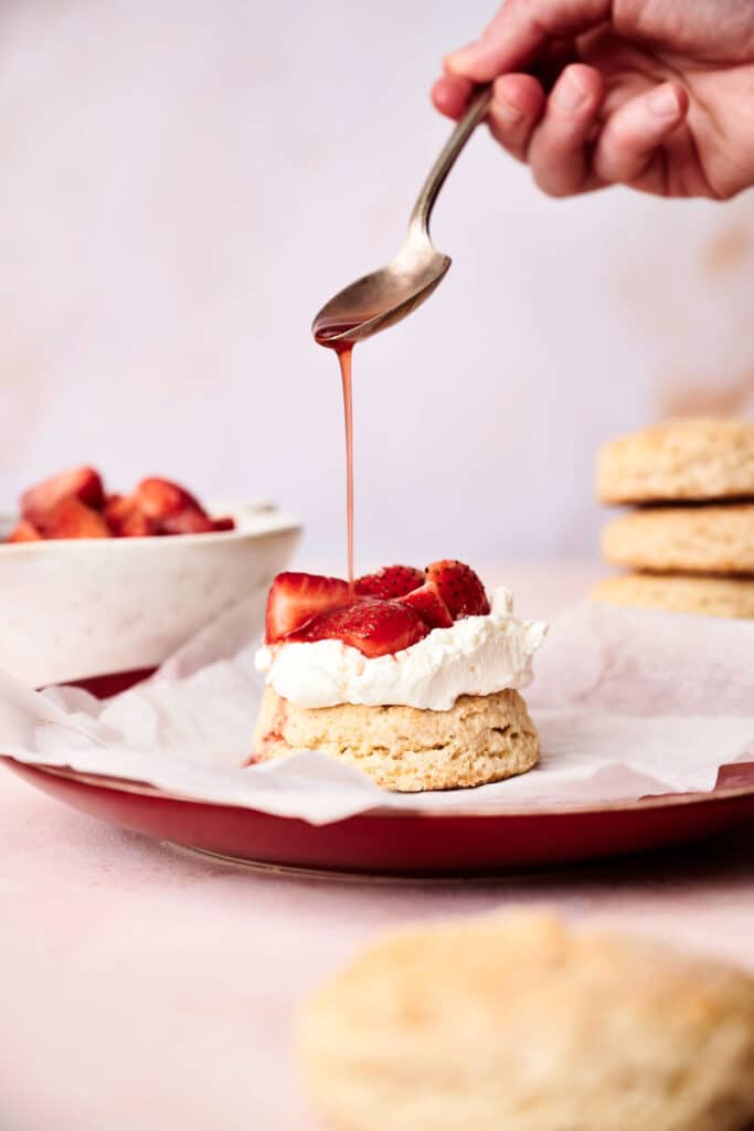 A spoon drizzles syrup over a biscuit topped with whipped cream and sliced strawberries on a red plate, reminiscent of a delicious strawberry shortcake. A bowl of additional strawberries and more biscuits are in the background.