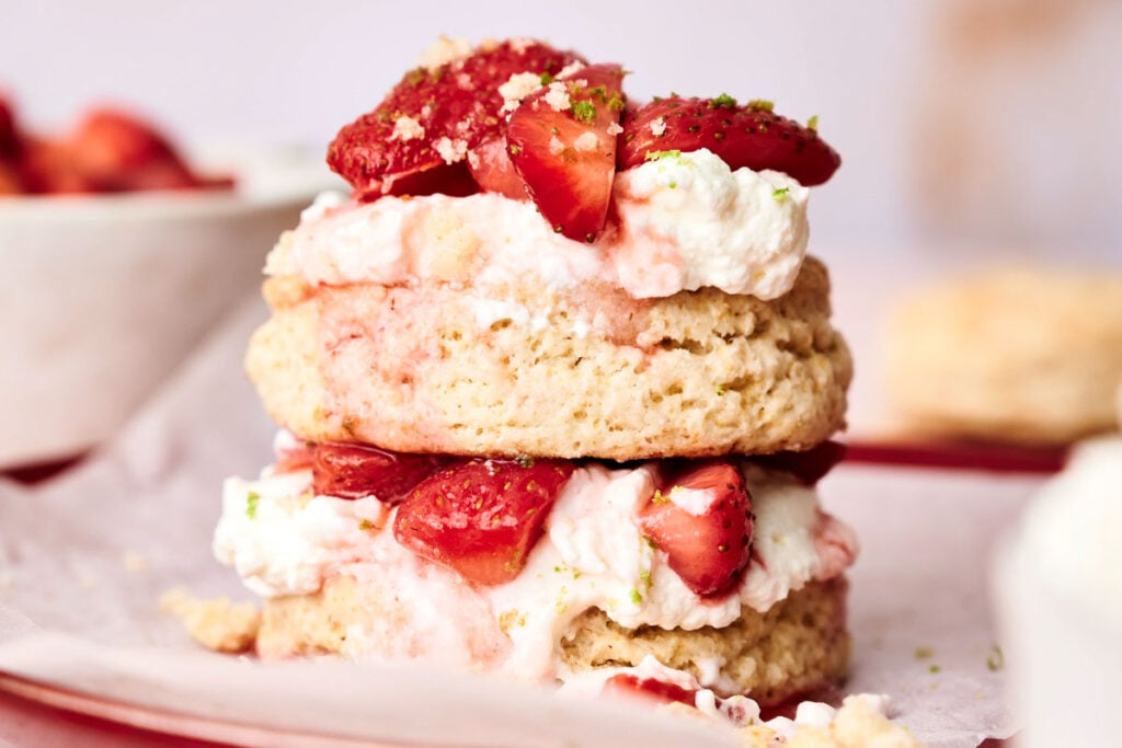 A close-up of a classic Strawberry Shortcake with two biscuit layers, whipped cream, and sliced strawberries on parchment paper.
