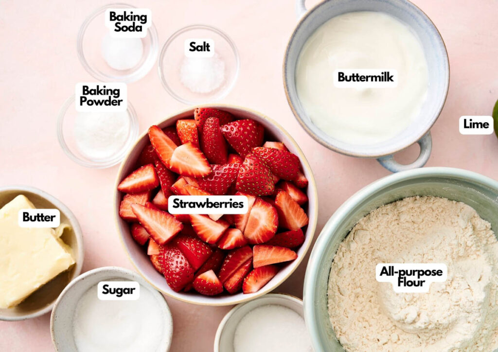 Bowls of ingredients for baking strawberry shortcake, including sliced strawberries, all-purpose flour, buttermilk, butter, sugar, baking powder, baking soda, salt, and a lime.