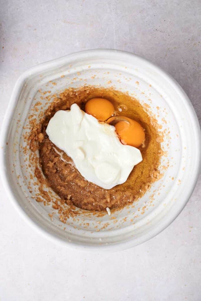 Mixing bowl containing brown sugar, two cracked eggs, and a dollop of yogurt.