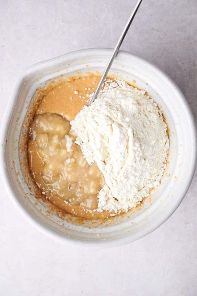 A mixing bowl containing a mixture of brown batter, mashed bananas, and a pile of flour, with a spoon partially visible in the bowl.