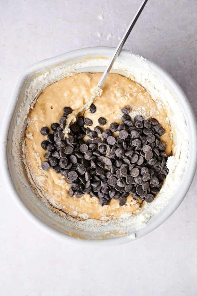 A mixing bowl with cookie dough and chocolate chips, with a spoon sticking out, sits on a countertop.