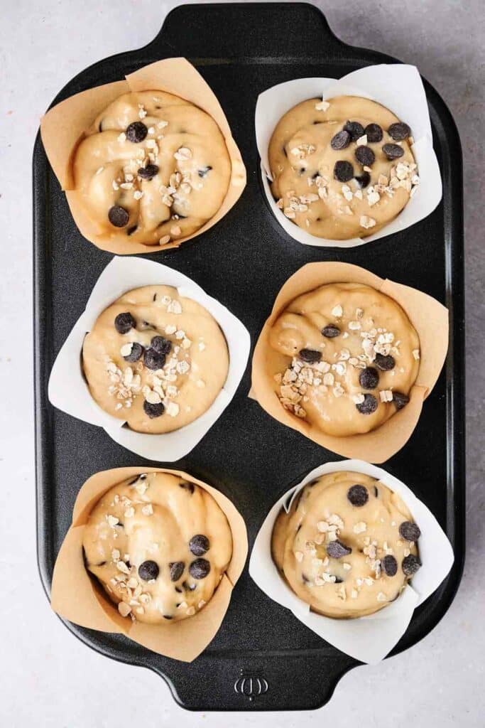 A muffin tray with six unbaked muffins lined with paper holders; the muffins are topped with chocolate chips and oats.