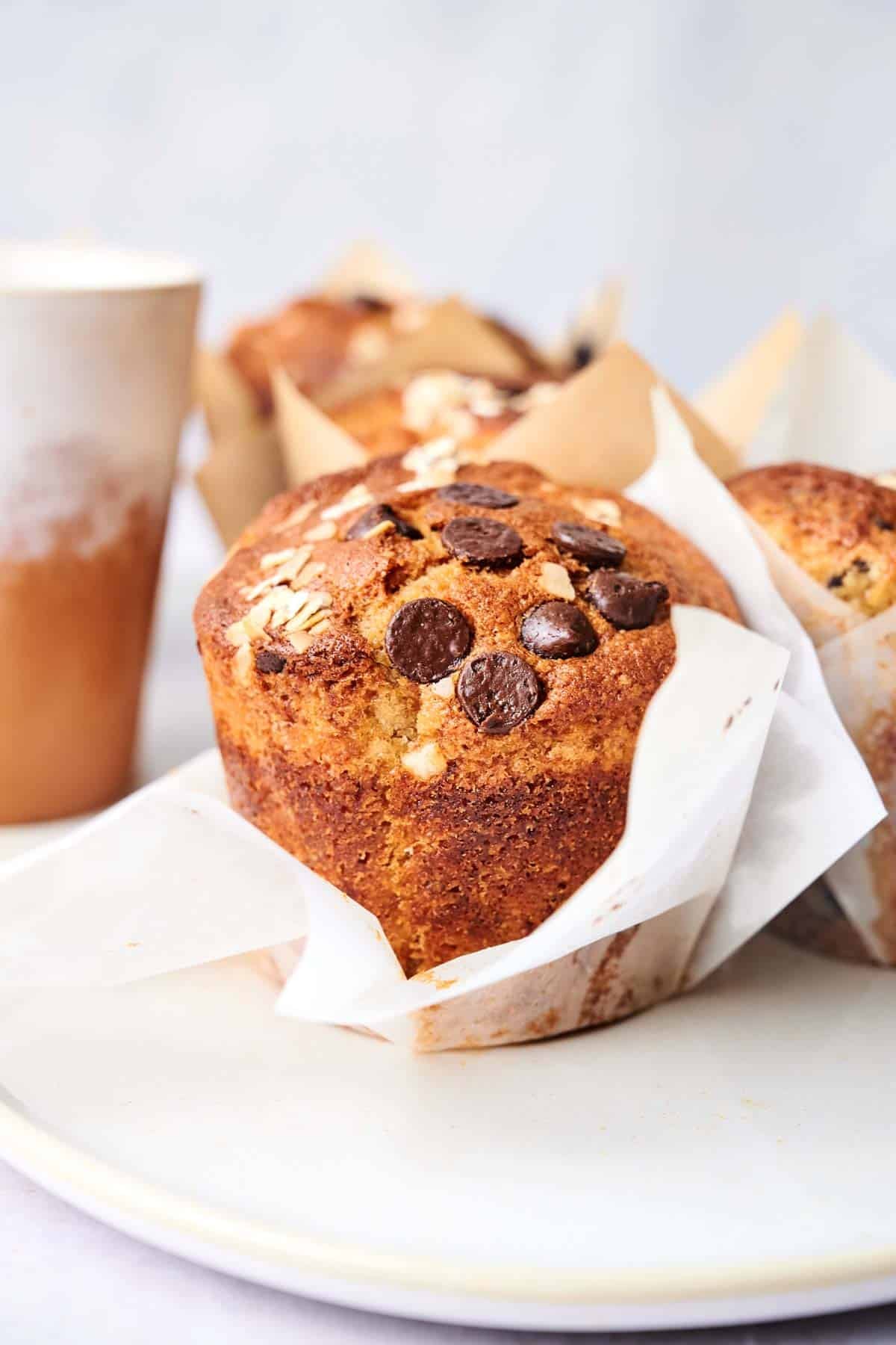 Close-up of a plate with banana chocolate chip muffins wrapped in parchment paper, topped with oats and chocolate chips, next to a ceramic cup.