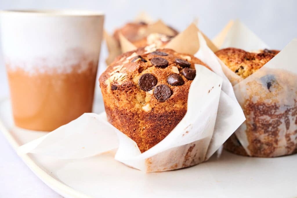 A close-up of a chocolate chip muffin wrapped in parchment paper, placed next to a ceramic cup on a white plate. Other muffins are blurred in the background.