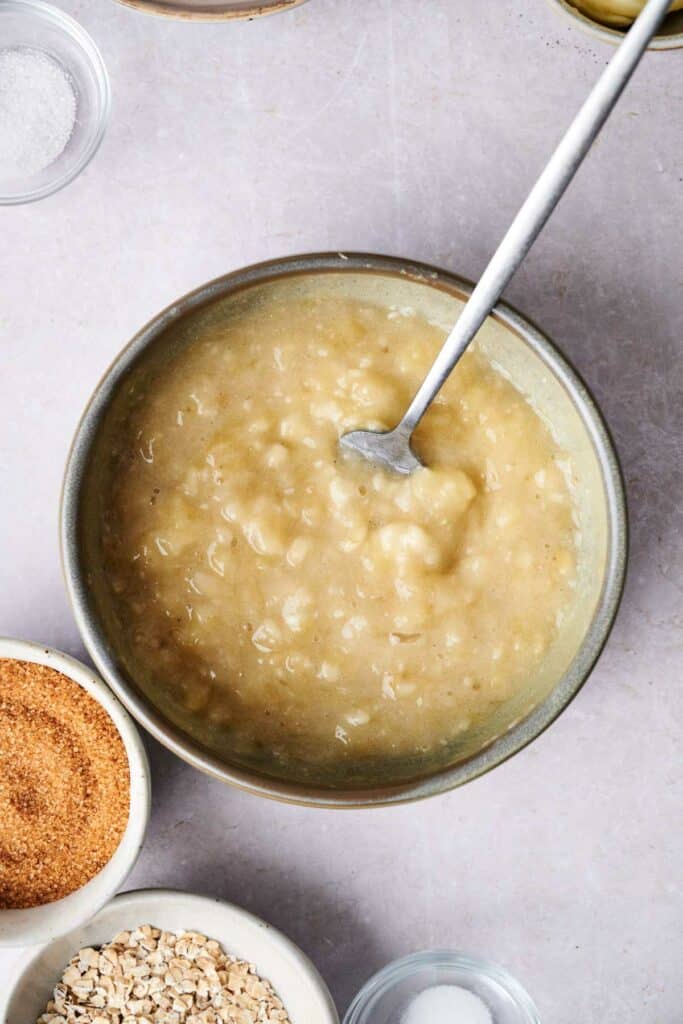 A bowl filled with mashed bananas and a spoon, surrounded by smaller bowls containing brown sugar, oats, and salt on a light surface.