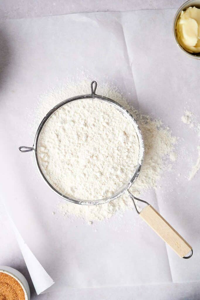 A sifter filled with flour is placed on a sheet of parchment paper. Various baking ingredients are partially visible around the edges of the image.