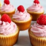 Lemon cupcakes with raspberry buttercream frosting, topped with fresh raspberries, are arranged in a delightful cluster on a pristine white surface.