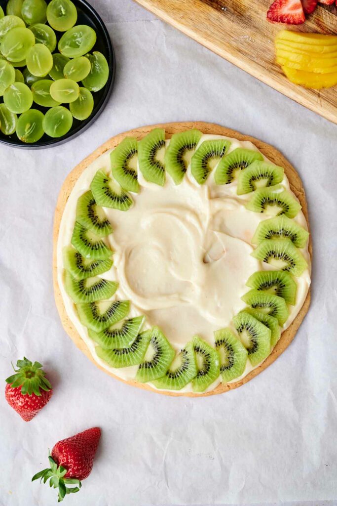 A round fruit pizza topped with a layer of cream and slices of kiwi arranged in a circle. Nearby are strawberries, a bowl of green grapes, and a wooden board with sliced mangoes and strawberries.
