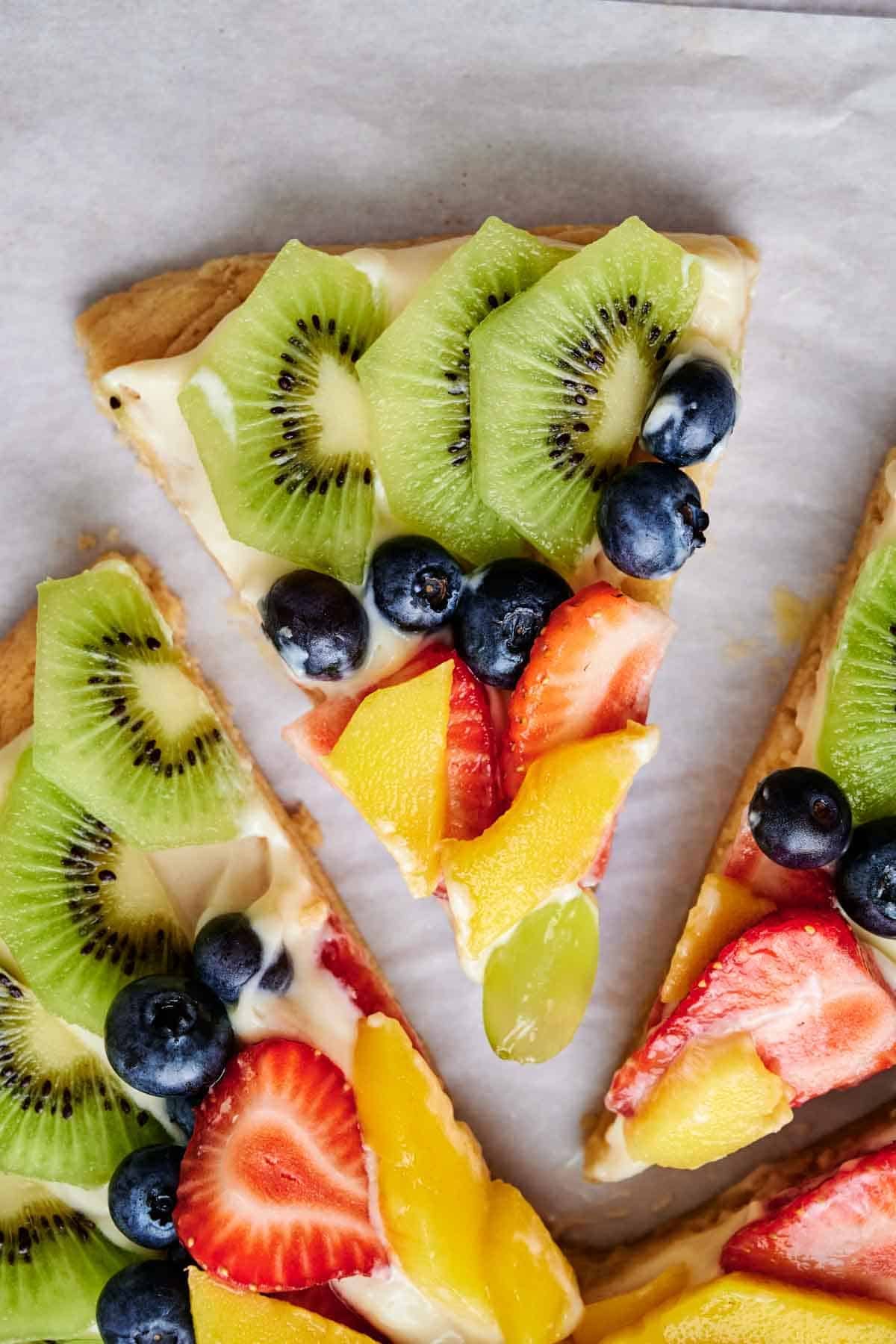 A slice of fruit pizza topped with kiwi, strawberries, blueberries, grapes, and mango on a parchment paper background.
