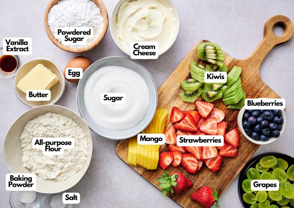Assorted baking ingredients in bowls and fresh fruits including kiwi, mango, strawberries, blueberries, and grapes arranged on a cutting board. Each ingredient is labeled.