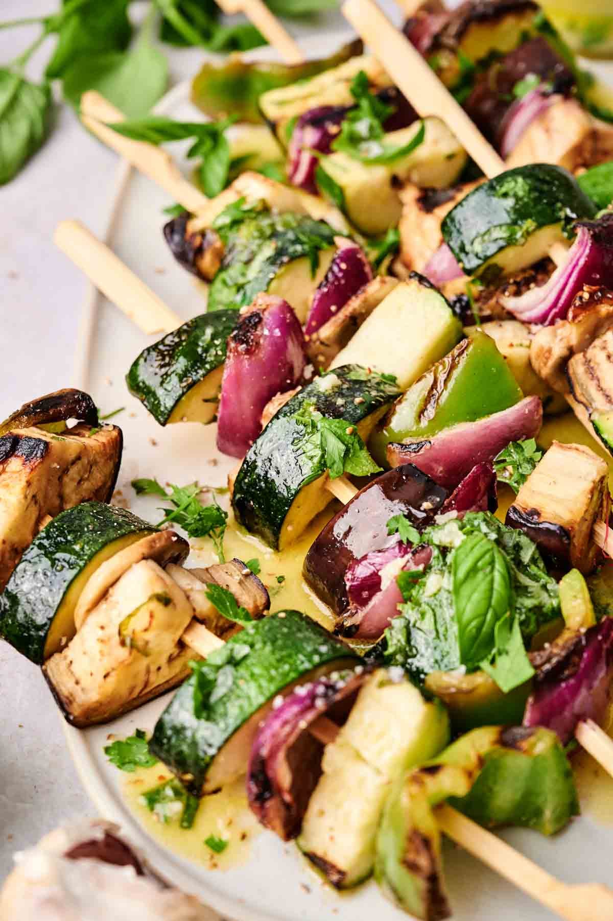 Close-up of grilled veggie skewers with zucchini, red onion, eggplant, and bell pepper pieces garnished with fresh herbs on a white plate.