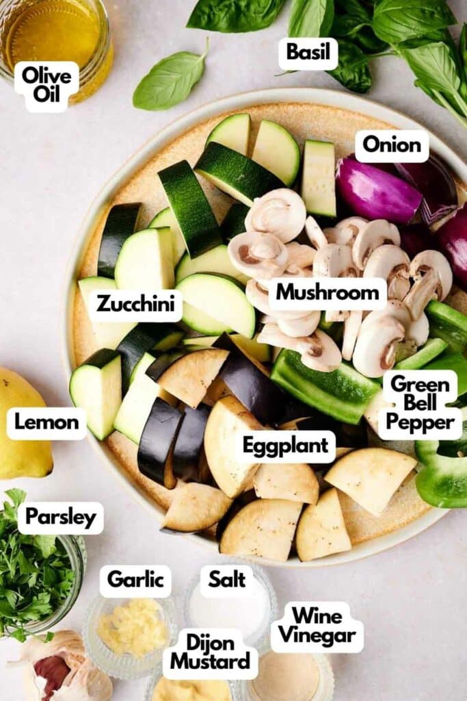 A plate with labeled chopped ingredients for grilled veggie skewers including zucchini, eggplant, mushrooms, green bell pepper, onion, basil, parsley, garlic, lemon, salt, olive oil, Dijon mustard, and white wine vinegar.