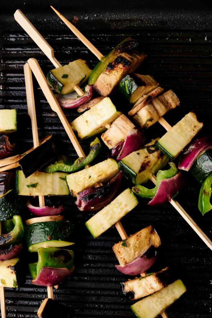 A close-up of grilled veggie skewers on a grill pan, featuring chunks of zucchini, eggplant, red onion, and green bell pepper.