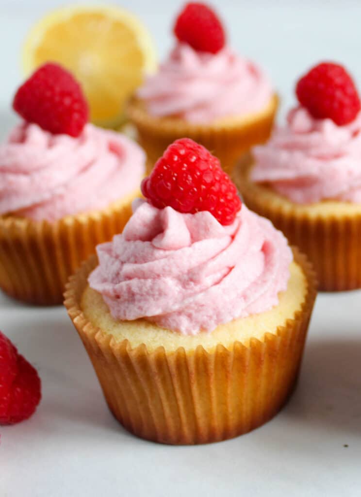 Lemon cupcakes with raspberry buttercream frosting are topped with pink frosting and a fresh raspberry, while a lemon slice accents the background.
