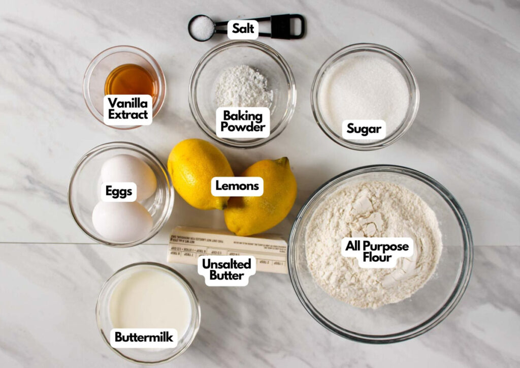 Baking ingredients on a marble surface, each labeled for Lemon Cupcakes with Raspberry Buttercream Frosting&mdash;lemons, eggs, buttermilk, unsalted butter, flour, sugar, baking powder, vanilla extract, and salt.