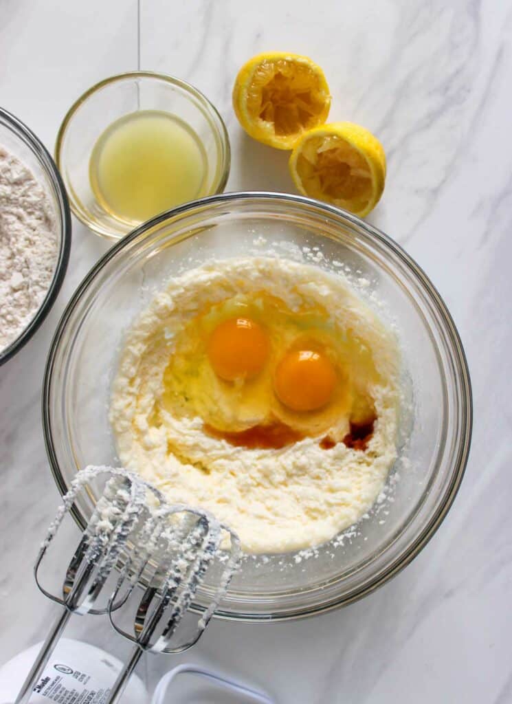 A mixing bowl containing two cracked eggs and other ingredients, with a hand mixer nearby, sets the stage for Lemon Cupcakes with Raspberry Buttercream Frosting. A bowl of lemon juice, flour, and two squeezed lemon halves lay ready on the marble countertop.