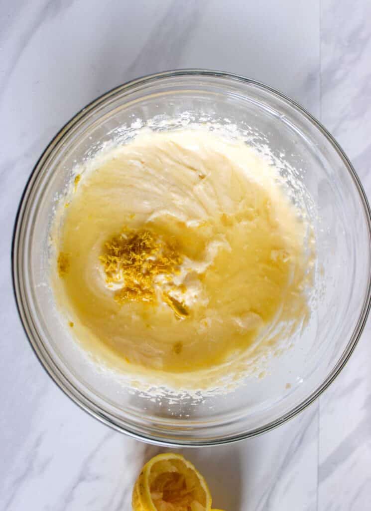 A glass bowl containing a mixture of beaten butter and sugar with lemon zest on top, perfect for making Lemon Cupcakes with Raspberry Buttercream Frosting. A halved lemon is positioned nearby on a white marble surface.