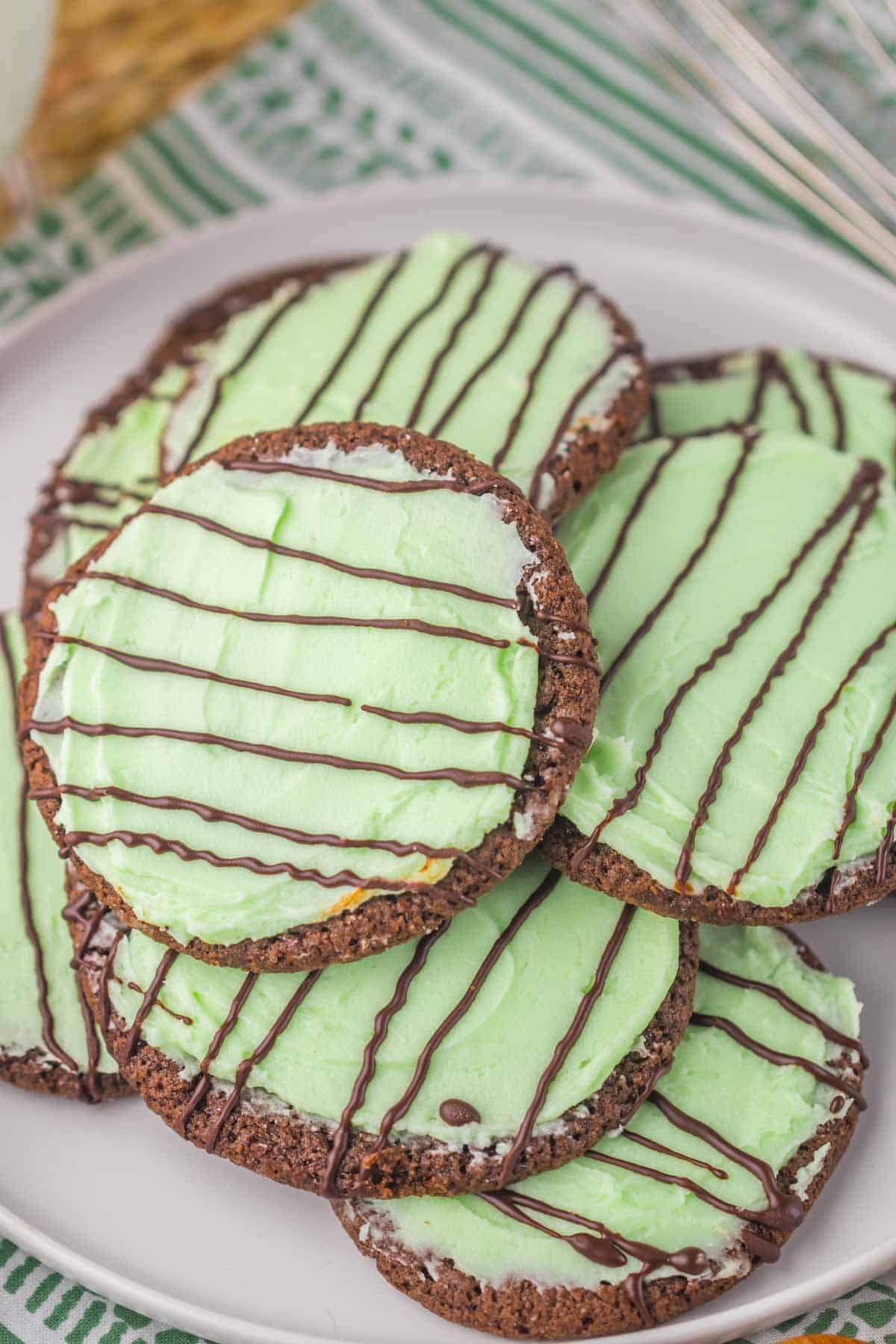 A plate of Thin Mint Chocolate Cookies with green frosting and chocolate drizzle, arranged on a white dish on a patterned tablecloth.