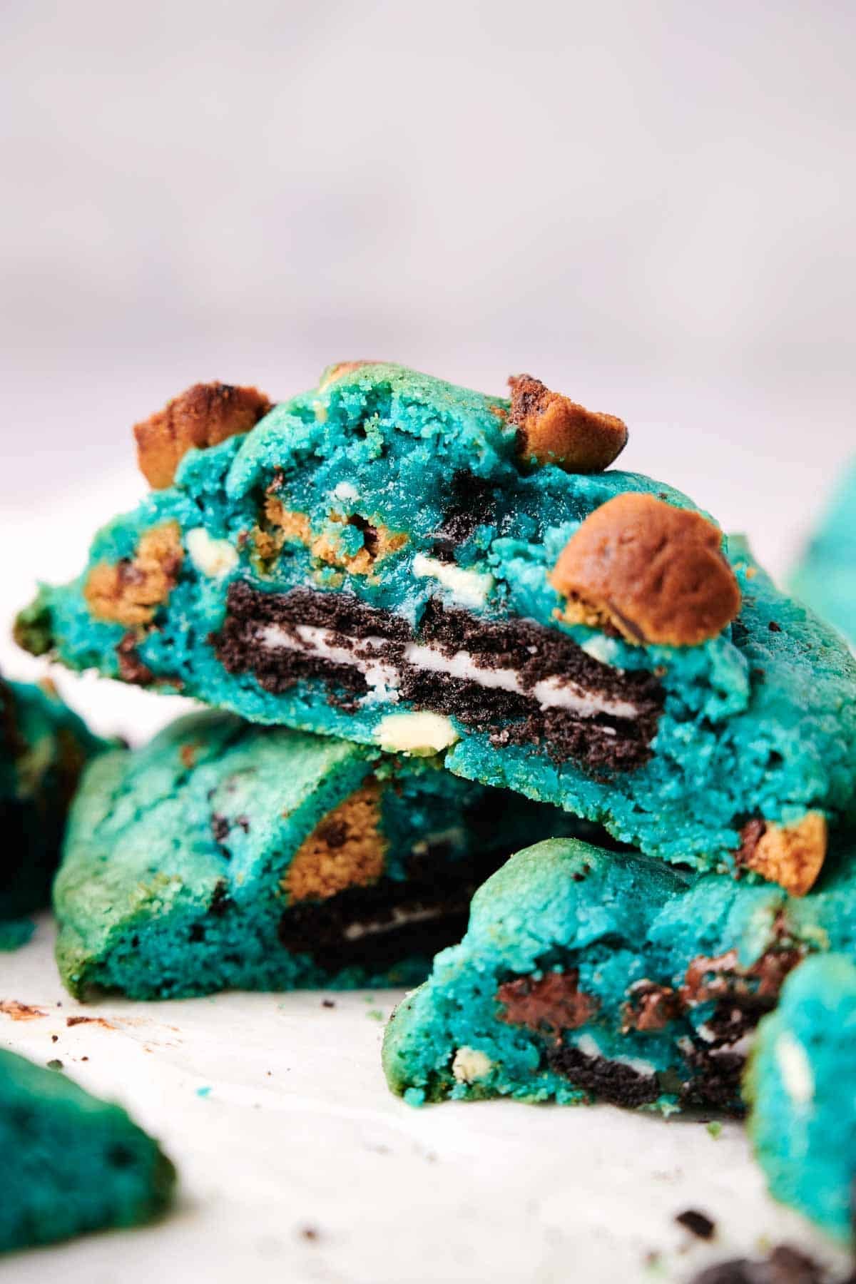 A close-up of a stack of vibrant blue Cookie Monster cookies, some broken to reveal a chocolate and cream center, located on a white surface. Small brown chunks are visible within the blue cookie dough.