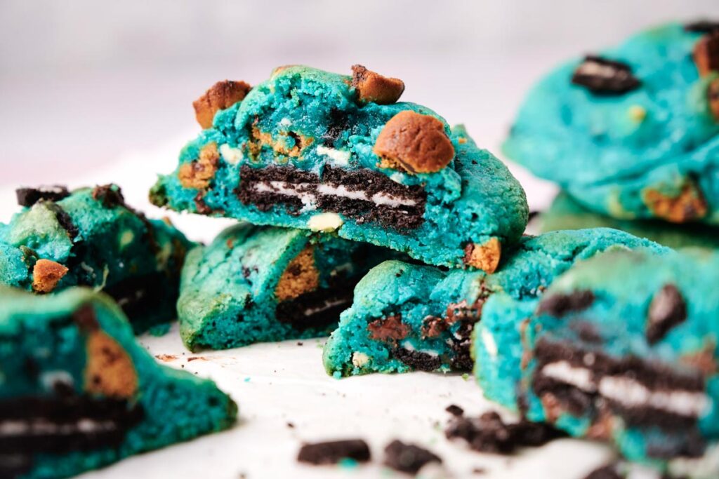 Bright blue Cookie Monster cookies are stacked and broken open, revealing an Oreo cookie and chocolate chip pieces inside. Crumbs are scattered on the surface.