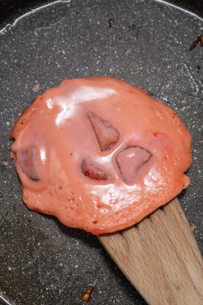 A piece of pink, melted candy with embedded fragments being lifted by a wooden spatula in a pan.