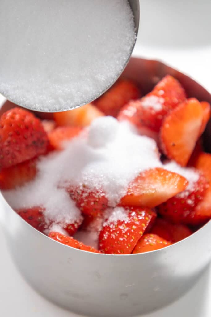 A metal measuring cup pouring granulated sugar onto a bowl of sliced strawberries.