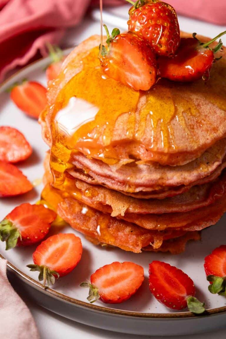A stack of pancakes topped with sliced strawberries and drizzled with syrup, served on a plate with additional halved strawberries around the edges.