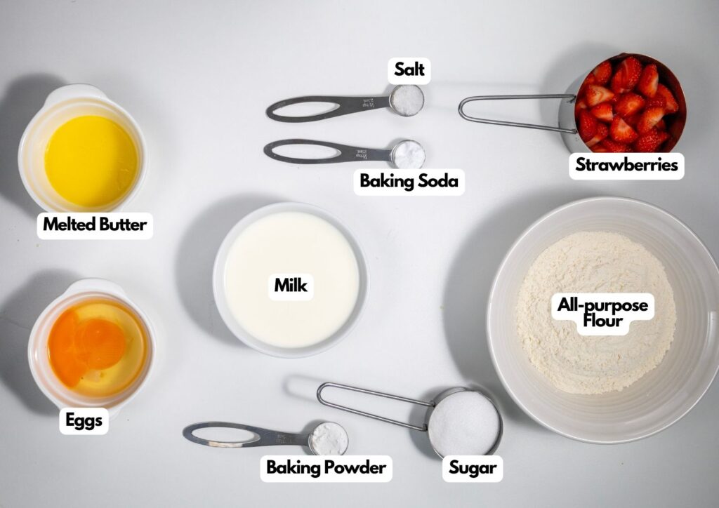 Top-down view of labeled baking ingredients in bowls and measuring cups: melted butter, eggs, milk, salt, baking soda, baking powder, sugar, all-purpose flour, and strawberries.