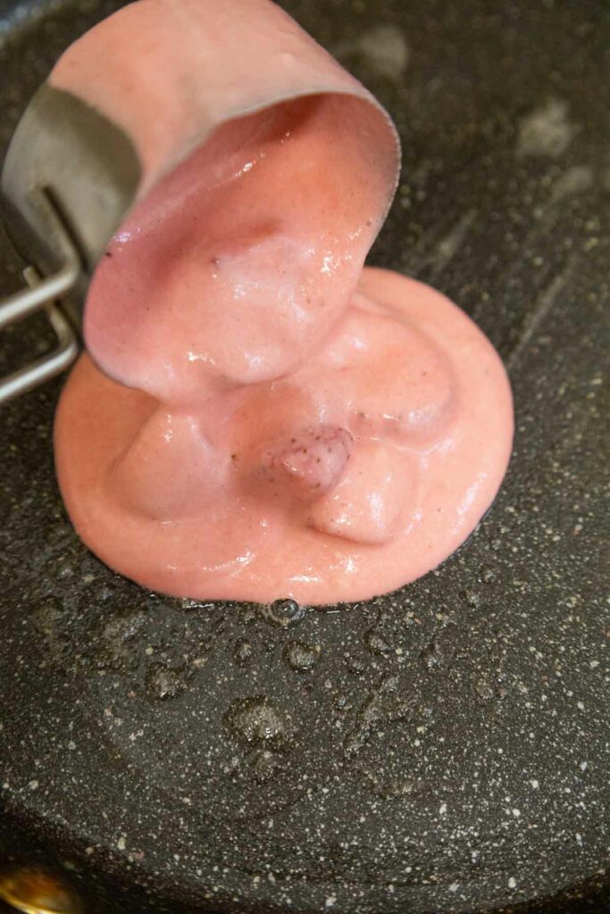 Thick pink batter being poured from a measuring cup onto a black griddle.