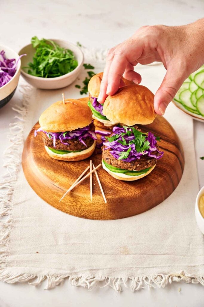 A hand adds a bun to a colorful Veggie Burger Slider with lettuce and purple cabbage, arranged on a wooden board with toothpicks and bowls of vegetables nearby.