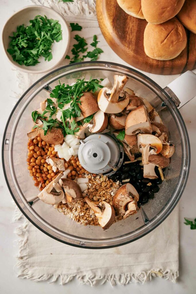 Food processor filled with raw mushrooms, beans, oats, and parsley, placed on a white cloth. A bowl of fresh parsley and a wooden board with bread rolls ready for making Air Fryer Veggie Burger Sliders are visible in the background.