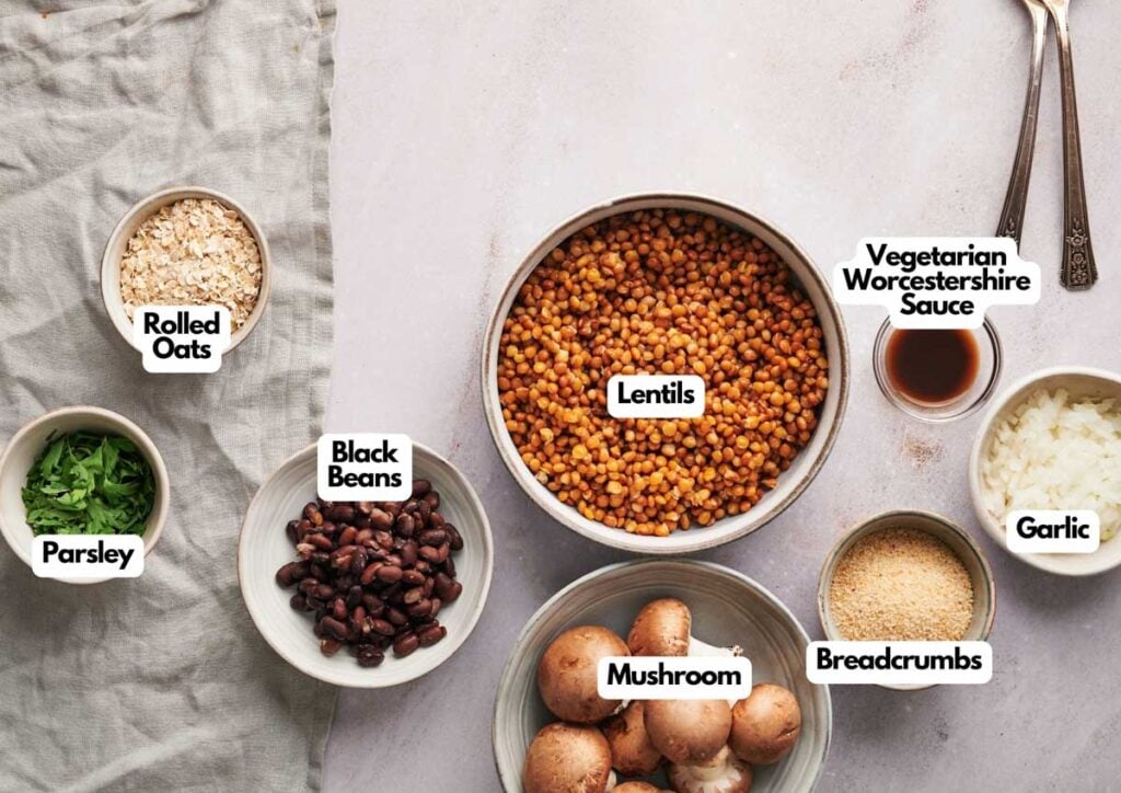 Bowls containing rolled oats, parsley, black beans, lentils, vegetarian Worcestershire sauce, garlic, breadcrumbs, and mushrooms—everything you need to craft flavorful Veggie Burger Sliders—arranged artfully on a white cloth.
