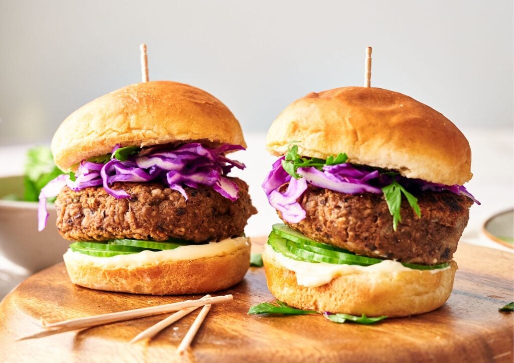 Two Veggie Burger Sliders on buns with cucumber slices, mayonnaise, purple cabbage, and parsley, displayed on a wooden board with toothpicks nearby.
