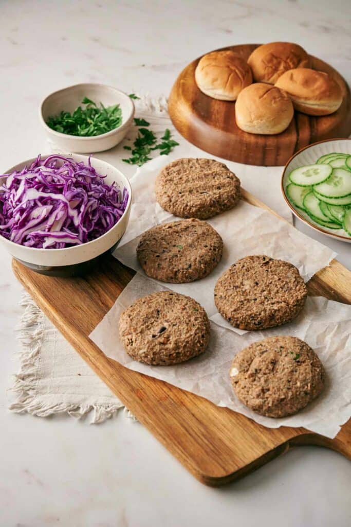 A wooden cutting board holds four plant-based veggie burger sliders on wax paper, accompanied by bowls of sliced cucumbers, shredded purple cabbage, and leafy greens. Sandwich buns can be seen in the background.