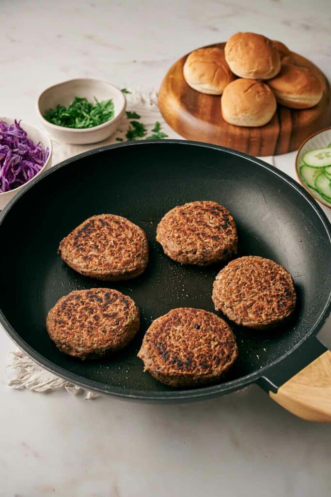 A pan containing four cooked veggie burger sliders. Nearby are burger buns, a bowl of chopped herbs, sliced cucumber, and shredded red cabbage on a kitchen counter.