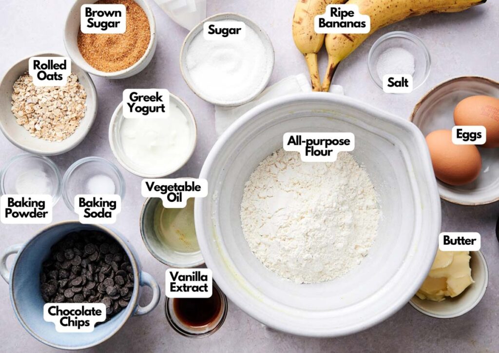 Assorted baking ingredients in bowls on a countertop, including flour, sugar, eggs, bananas, oats, butter, oil, yogurt, chocolate chips, baking powder, baking soda, salt, and vanilla extract—everything you need to make delicious Banana Chocolate Chip Muffins.