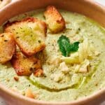 A bowl of creamy soup topped with crispy croutons, shredded cheese, a mint sprig, and a drizzle of olive oil.