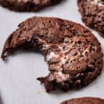 Close-up of a chocolate cookie with a large bite taken out of it, revealing a gooey inner filling on a white background. Other marshmallow swirl cookies can be seen in the blurry background.