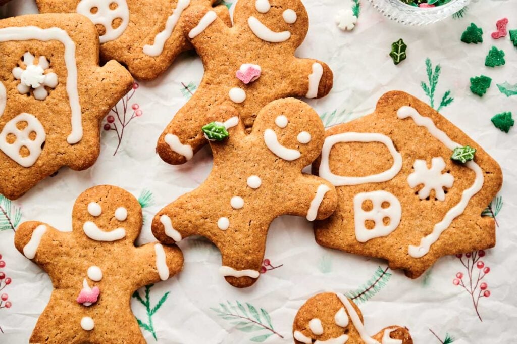 Several decorated Gingerbread Cookies, including gingerbread people and houses, are arranged on festive paper with holiday-themed icing and sprinkles.
