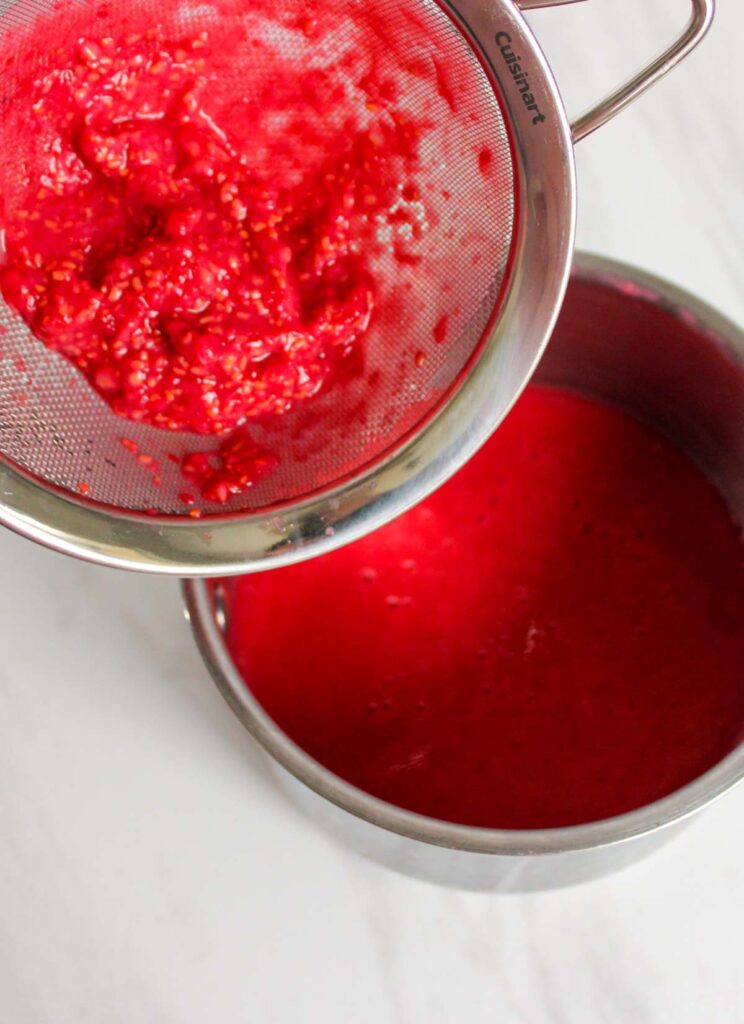 A metal strainer holds mashed raspberries above a pot filled with strained raspberry liquid on a white surface, capturing the vibrant essence perfect for Lemon Cupcakes with Raspberry Buttercream Frosting.