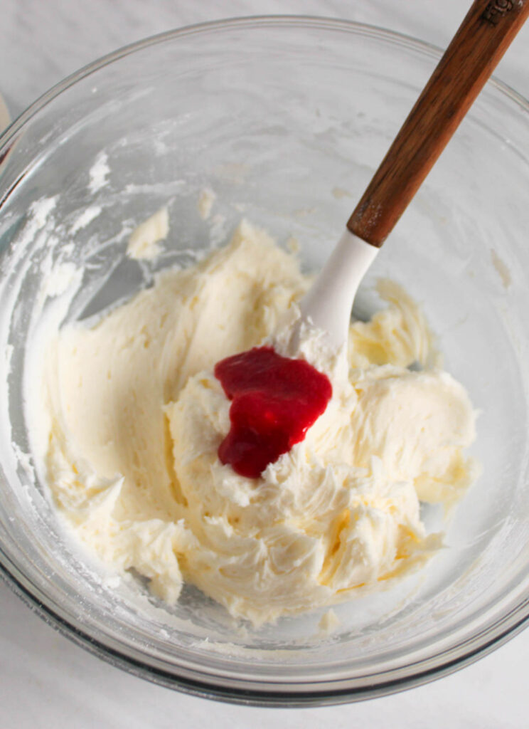 A glass bowl with whipped cream cheese mixture and a dollop of red fruit puree, reminiscent of the vibrant flavors in Lemon Cupcakes with Raspberry Buttercream Frosting, stirred with a white spatula with a wooden handle.