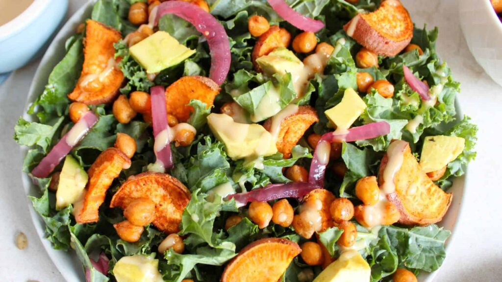 A salad topped with roasted sweet potatoes, chickpeas, avocado, kale, red onions, and a drizzle of dressing.