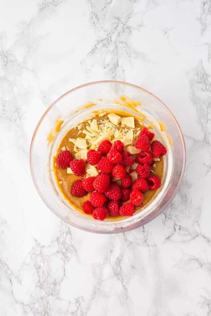 A glass bowl contains a mixture of brown batter, white chopped chocolate, and fresh raspberries on a marble surface, the perfect beginning for Raspberry and White Chocolate Blondies.