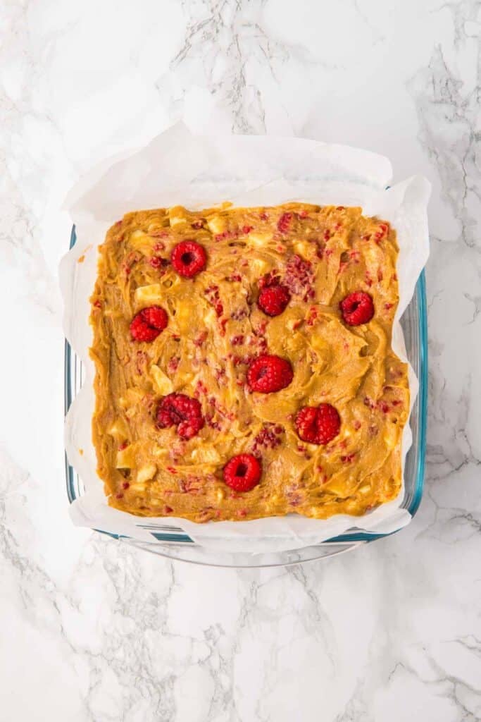 A square pan with parchment paper holds unbaked raspberry and white chocolate blondies batter, topped with fresh raspberries. The pan rests on a marble countertop.