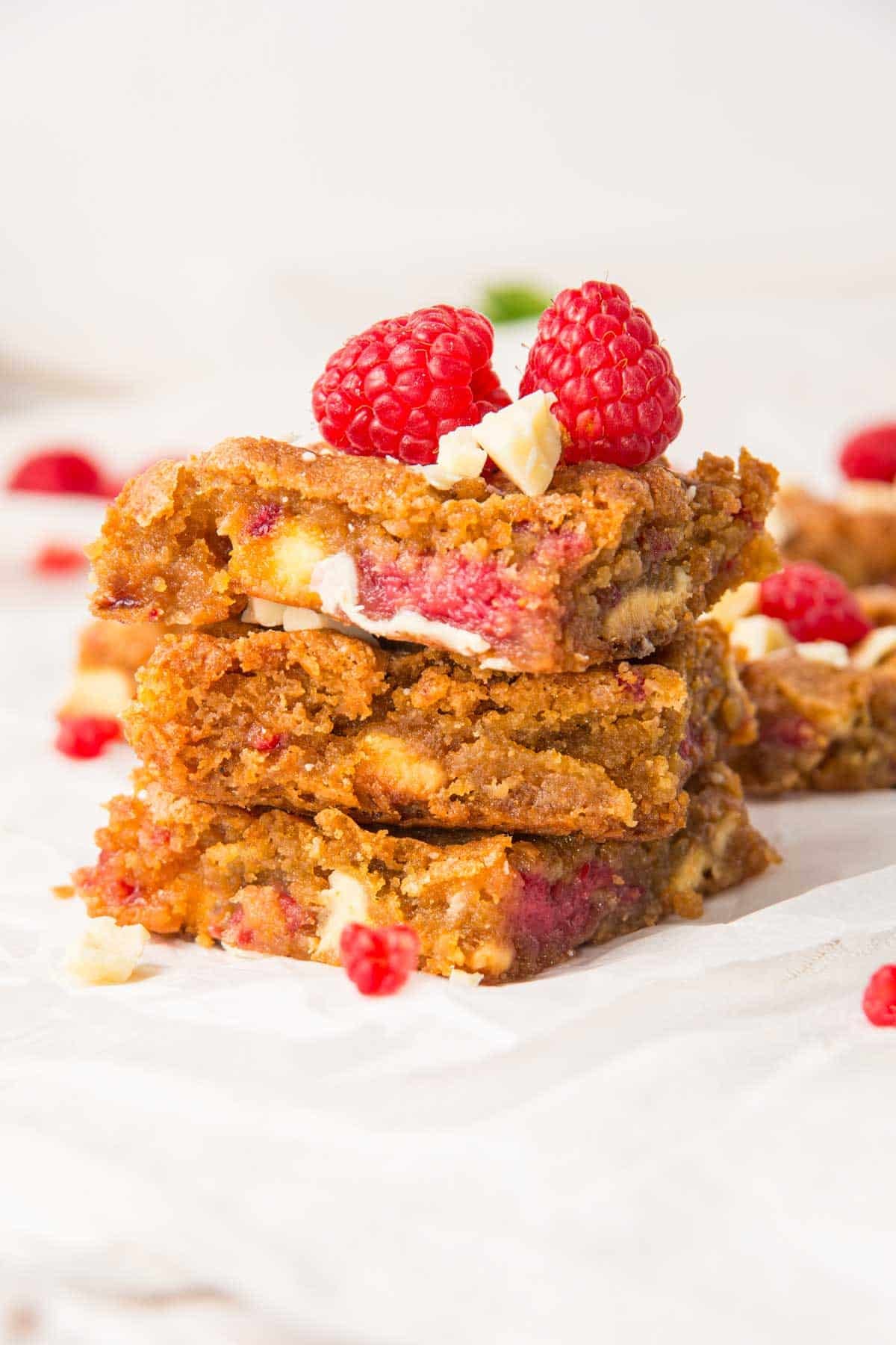 A tantalizing stack of Raspberry and White Chocolate Blondies garnished with fresh raspberries and white chocolate chunks sits on a pristine white surface. More raspberries and blondies are artfully scattered in the background, enhancing the irresistible charm.