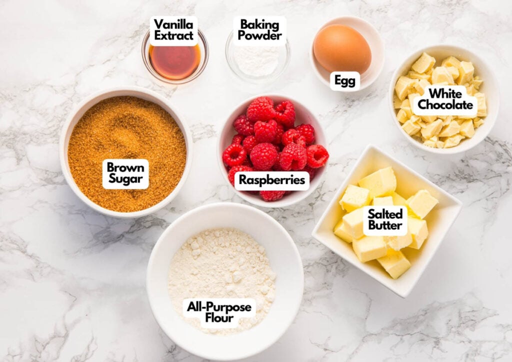 Top-down view of baking ingredients for Raspberry and White Chocolate Blondies in bowls on a marble surface, including brown sugar, all-purpose flour, raspberries, vanilla extract, baking powder, egg, white chocolate, and salted butter.