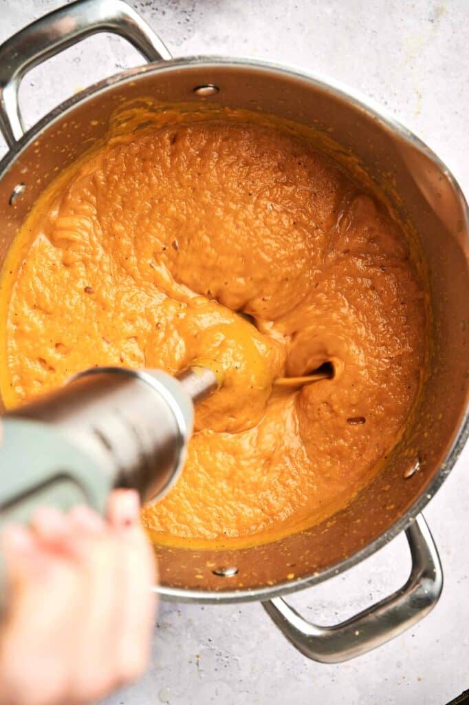 A person uses an immersion blender to puree a thick orange mixture in a large metal pot, creating a smooth and creamy butternut squash soup.