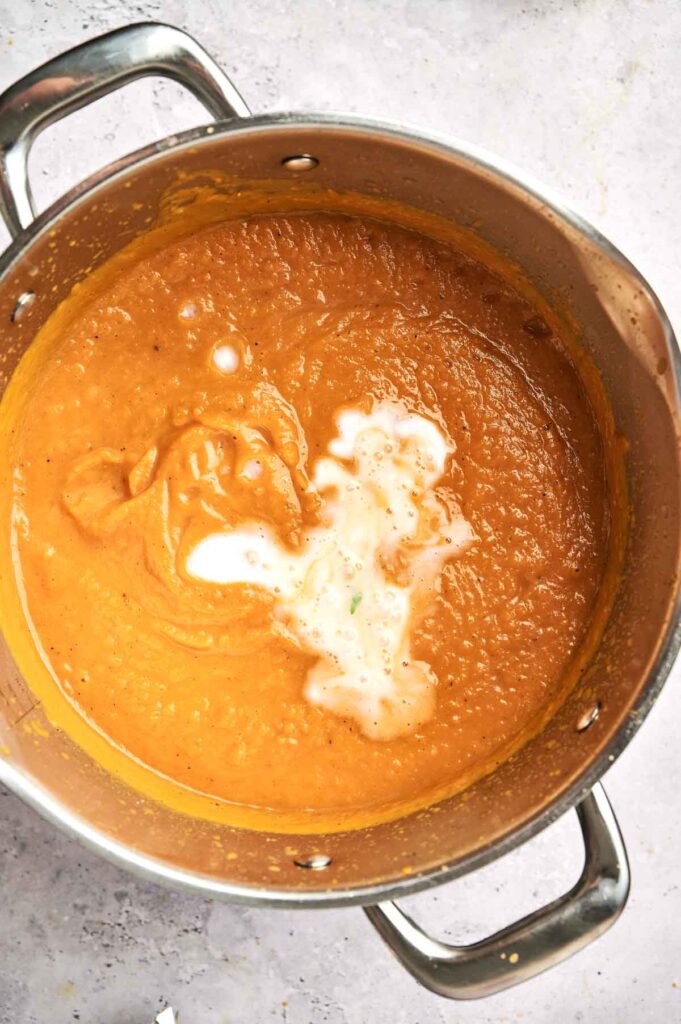 A stainless steel pot contains creamy butternut squash soup with a swirl of white cream on top. The background is a light grey surface.