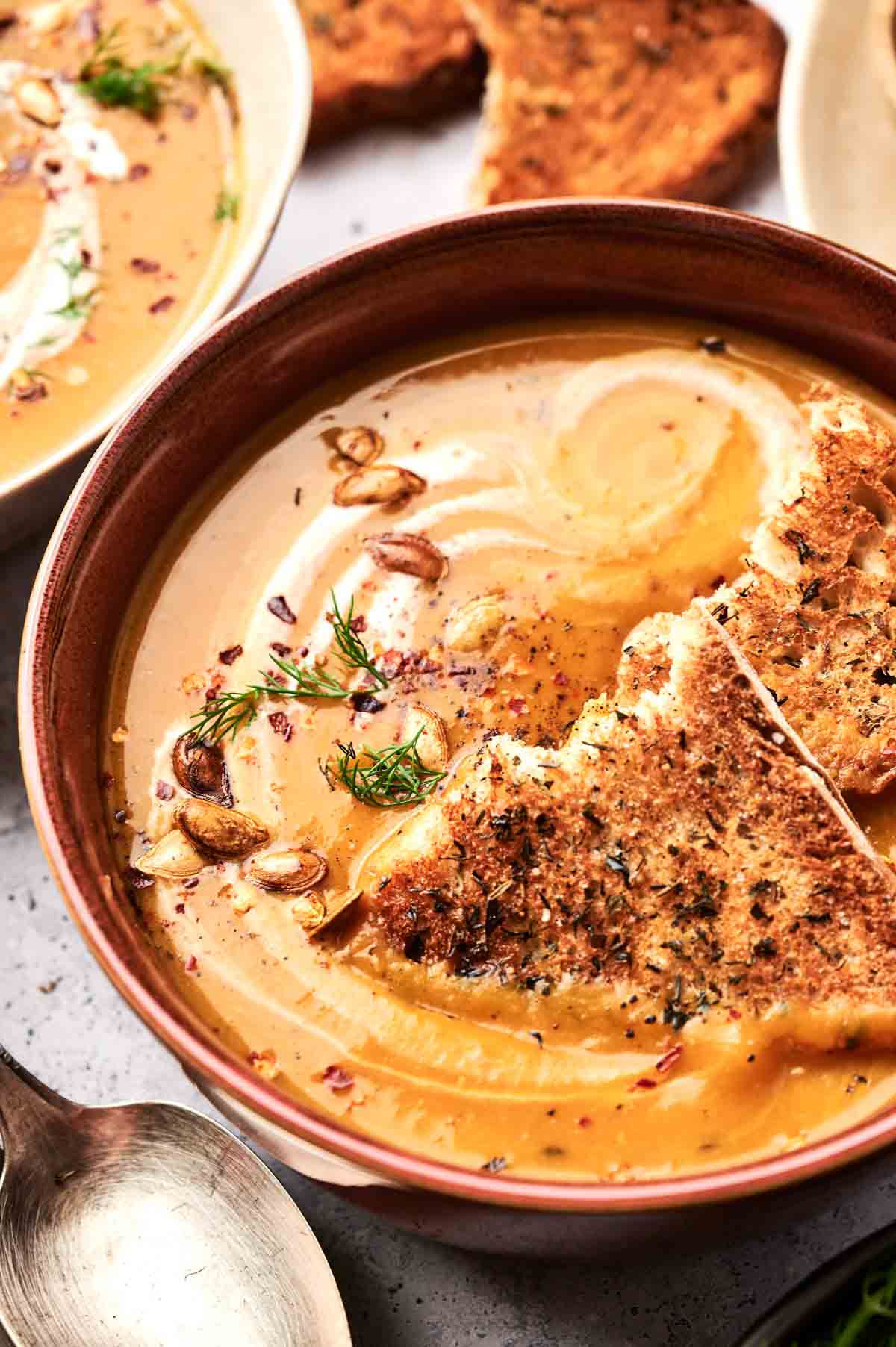 A bowl of creamy butternut squash soup garnished with herbs and seeds, served with a slice of toasted bread. A spoon and additional pieces of toast are visible in the background.